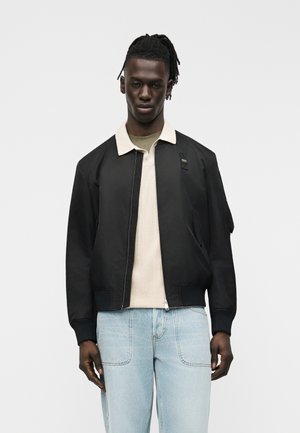 Homme aux dreadlocks portant une veste bomber noire sur une chemise beige clair et un jean bleu clair, debout devant un fond uni.