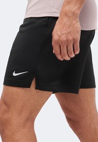 Άνδρας με μαύρα σορτς Nike και μπεζ τοπ, που δείχνει πλάγια όψη των σορτς και το χέρι του άνδρα να ακουμπά κοντά στον μηρό.