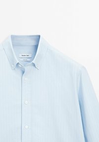 Light blue button-up shirt made of fabric with vertical white stripes, featuring a traditional collar and white buttons. Label visible inside.