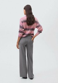 Donna con lunghi capelli scuri che indossa un maglione a righe rosa e grigio e pantaloni grigi a palazzo, in piedi con la schiena rivolta e una mano in tasca.