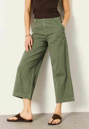 Vrouw draagt olijfgroene wijde cropped broek met bloemenborduursel en een bruine mouwloze top, staand met één hand in de zak.