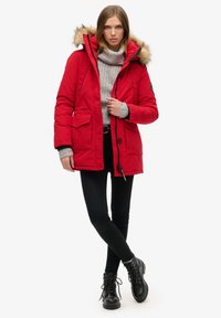 Red padded parka jacket with a fur-lined hood, featuring two front pockets, a zipper and button closure, and black accents. Worn over a gray sweater.