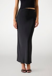 Schwarzer Satin-Midirock mit elegantem Design, das einen subtilen Glanz und einen seitlichen Schlitz aufweist, kombiniert mit schwarzen Riemchen-Heels.