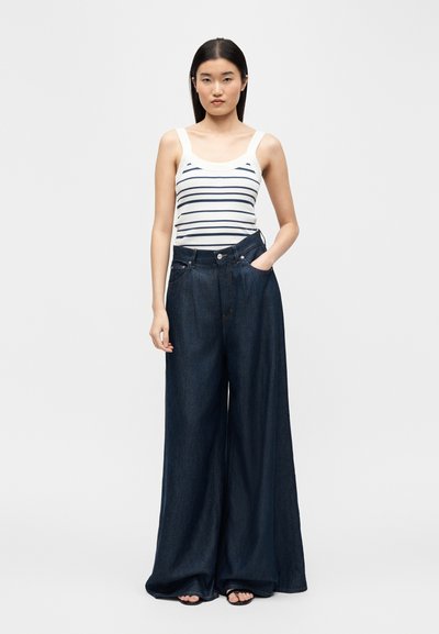 Young woman with straight dark hair wearing a white tank top with navy stripes and dark wide-leg denim jeans with one hand in pocket.