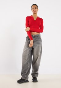 Rotes, geripptes Longsleeve-Oberteil mit V-Ausschnitt und Kragen, kombiniert mit locker sitzenden grauen Jeans und schwarzen Sneakern. Einfarbiger weißer Hintergrund.