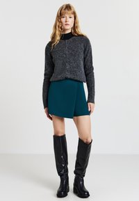 Donna con lunghi capelli biondi che indossa un maglione grigio scuro con bottoni, una gonna asimmetrica teal e stivali neri fino al ginocchio, in piedi su uno sfondo bianco.