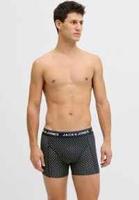 Jauns vīrietis stāv krekla bez, valkājot melnas bokseršortus ar balamu palmu koku rakstu un "JACK & JONES" jostas joslu, skatoties pa labi.