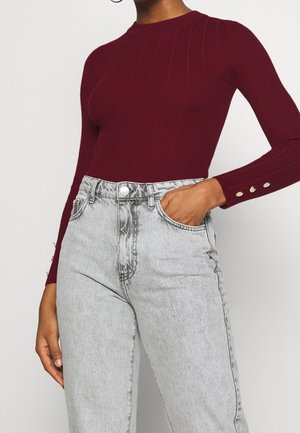 Femme portant un haut bordeaux ajusté à manches longues avec des boutons aux poignets et un jean gris clair taille haute, main dans la poche.