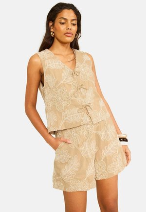 Woman wearing beige sleeveless top and matching shorts with white floral embroidery, accessorized with gold hoop earrings and bangles.