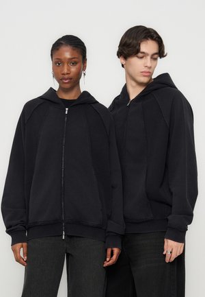 Twee jonge volwassenen dragen oversized zwarte hoodies met rits en staan zij aan zij tegen een effen witte achtergrond.