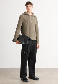 Maslinasto zeleni rebrasti pulover s polu-zip ovratnikom, uparen s crnim cargo hlačama koje imaju bočne džepove i srebrne detalje, držane s crnom clutch torbicom.