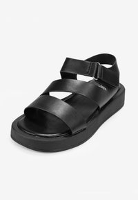 Sandalia de cuero negra con tiras cruzadas, hebilla ajustable y suela gruesa. Textura suave con un detalle de costura mínimo.