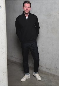 Sudadera negra con cremallera, vaqueros azul oscuro y zapatillas de color claro. Tela suave con un corte relajado, de pie contra una pared de hormigón.