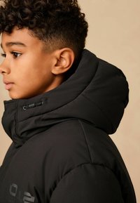 Čierna izolovaná bunda s vysokým golierom a kapucňou so sťahovacou šnúrkou. Mäkkučký, hladký materiál s prešívanými detailmi a logom na prednej strane.
