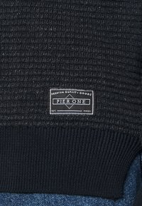 Dunkel strukturierter Pullover mit geripptem Saum, versehen mit einem schwarz-weißen Markenschild, auf dem "Pier One" steht, gegründet im Jahr 2011.