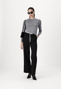 Graues Langarmshirt, schwarze weit geschnittene Hose mit weißem Kordelzug, schwarze Tasche und Sonnenbrille. Glatte Textur, figurbetontes Oberteil und verlängernde Silhouette.