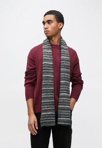 Burgundowy sweter z dzianiny z prążkowanym dekoltem, zestawiony z szarym paskowanym szalikiem z fakturowanej tkaniny. Model stoi na jednolitym tle.