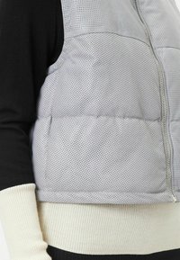 Graue gesteppte Weste mit perforierter Textur und silbernem Reißverschluss, die über einem schwarzen Ärmel und einem cremefarbenen gerippten Bund getragen wird.