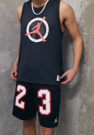 Osoba ubrana w czarną koszulkę i krótkie spodenki do koszykówki z czerwono-białym numerem 23 i czerwonym logo Jumpman, stojąca przy szarej betonowej ścianie.