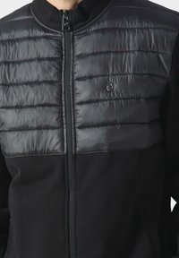 Chaqueta acolchada negra con una parte superior lisa y brillante y tela texturizada en la parte inferior. Cierre con cremallera y cuello alto.