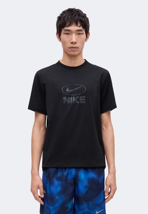 Jeune homme portant un t-shirt noir Nike et un short bleu à motifs, debout droit devant un fond clair uni.