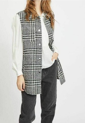 Kvinde iført en sort-hvid tjekket ærmeløs frakke over en hvid langærmet bluse og sorte jeans.
