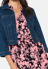 Jeansjacka med långa ärmar, ljusblå färg, två framfickor. Nedanför en rosa blommig klänning på mörk bakgrund, med dragsko i midjan.