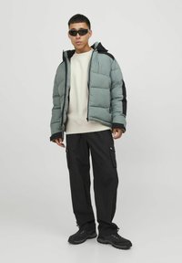 Padded green jacket with a black hood and accents, beige sweater, black cargo pants, and black shoes, worn by a person in a standing pose.