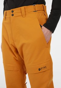 Oranje sneeuwbroeken van duurzame materialen, met een riem in de taille, twee voorkiethoeken en een logo op het onderbeen. Soepele textuur.