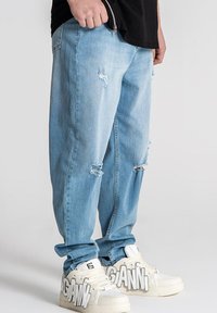 Jeans desgastados de color azul claro con rotos en las rodillas, combinados con zapatillas blancas que tienen texto en negrita en el lateral. Ajuste suelto a medida.