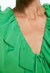 Nahaufnahme des Halses und oberen Brustbereichs einer Person, die eine leuchtend grüne Bluse mit Rüsche am Kragen und einem kleinen Bindeband am Ausschnitt trägt.