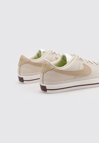 Zapatillas beige con suelas blancas y detalles en marrón, con una parte superior texturizada y un forro verde. Logo grabado en el talón.