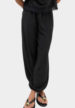 Person iført løse sorte bukser med elastiske manchetter, en sort blonder top og sorte sandaler med gyldne dekorative detaljer.