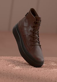 Brązowe skórzane trampki w stylu high-top z okrągłym noskiem, bocznymi paskami i czarną gumową podeszwą. Posiadają sznurowany design i tylne uchwyty.