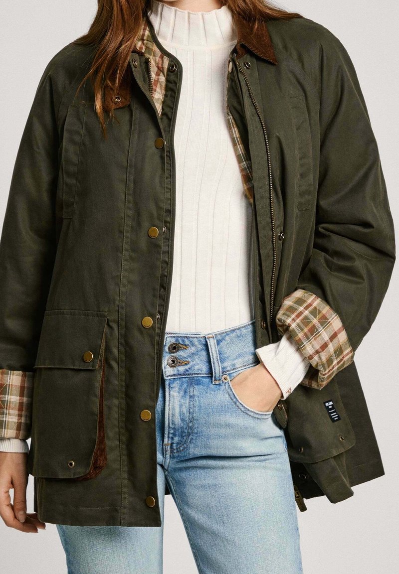 Olivgrön jacka med foder i flanell, knappstängning och två framfickor. Bärs över en vit ribbad tröja och blå jeans.