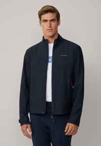 Giacca softshell blu navy con colletto alto, chiusura con zip e logo rosso a contrasto. Presenta una vestibilità rilassata e una superficie liscia.