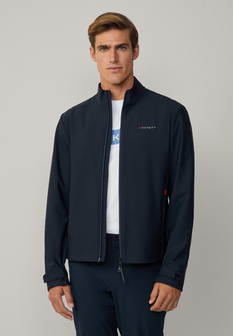 Giacca softshell blu navy con colletto alto, chiusura con zip e logo rosso a contrasto. Presenta una vestibilità rilassata e una superficie liscia.