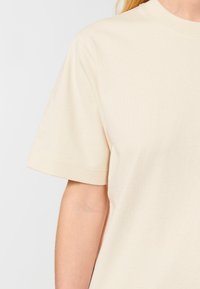 Kortærmet t-shirt i lys beige bomuld med rund halsudskæring, glat tekstur og ren syning langs ærmerne og kanten.