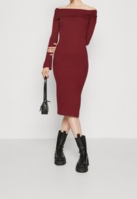 Robe à manches longues en maille côtelée bordeaux avec un décolleté bardot. Associée à un petit sac à bandoulière noir et des bottines noires épaisses.