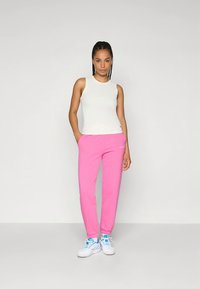 Biała bluzka bez rękawów w zestawieniu z różowymi dresami. Spodnie mają elastyczne mankiety i logo. Buty sportowe są białe z niebieskimi akcentami.