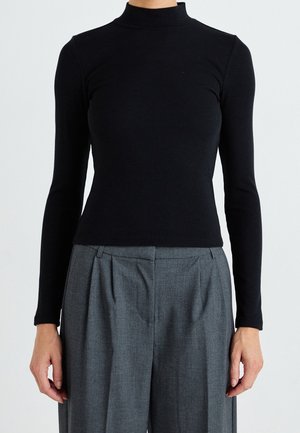 Persoon die een aansluitend zwart coltruitje met lange mouwen en hoge grijze geplooide broek draagt tegen een effen achtergrond.