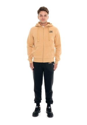 Un hombre posa con una sudadera con capucha beige y cremallera, pantalones deportivos negros y zapatillas negras sobre un fondo blanco.