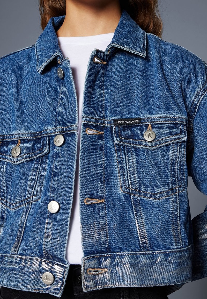 Vybledlá modrá džínová bunda s knoflíkovým zapínáním vpředu, dvěma náprsními kapsami a štítkem "Calvin Klein Jeans" na levé straně. Stříbrné kovové detaily.