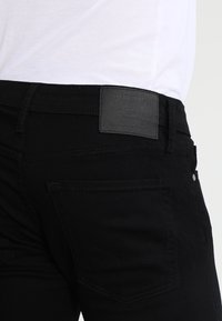 Svarta denimjeans med en slät yta, synliga sömmar och en svart läderetikett på midjan. Har standard bakfickor.