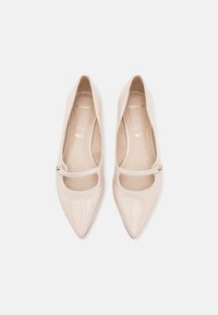 Une paire de ballerines Mary Jane beiges en cuir verni à bout pointu avec des brides simples et des accents en boutons dorés, présentées de dessus sur fond blanc.