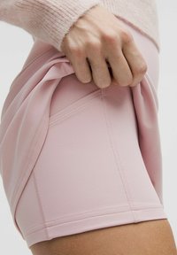 Svijetloružičaste atletske kratke hlače izrađene od rastezljivog materijala s vidljivim detaljima šavova; ručna hvata materijal pokazuje fleksibilnost i prianjanje.