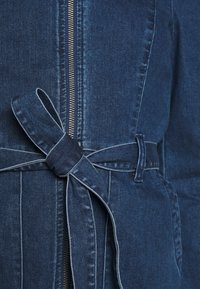 Veste en denim bleu foncé avec une fermeture éclair argentée centrale et une ceinture en tissu nouée en nœud à la taille.