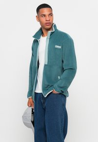 Teal fleecejakke med høy krage, glidelås foran og kontrasterende lysegrønne detaljer. Har to frontlommer og et logo-patch.