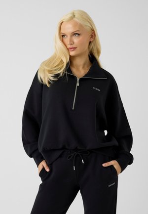 Donna bionda che indossa una felpa nera con zip Guess e pantaloni della tuta neri coordinati, con le mani nelle tasche, posando davanti a uno sfondo neutro.