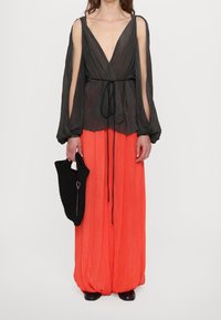 Haut noir plissé à manches longues avec un décolleté en V profond, associé à un pantalon large plissé orange vif et un sac tissé noir texturé.